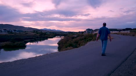 Reifer-Mann,-Der-Während-Eines-Erstaunlichen-Sonnenuntergangs-Auf-Einem-Fußweg-Neben-Einem-Fluss-Spaziert---Weg-Von-Der-Statischen-Kamera