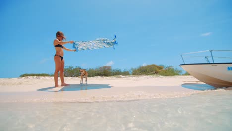 Girl-holding-beach-towel-over-above-dog-on-beach-near-a-boat