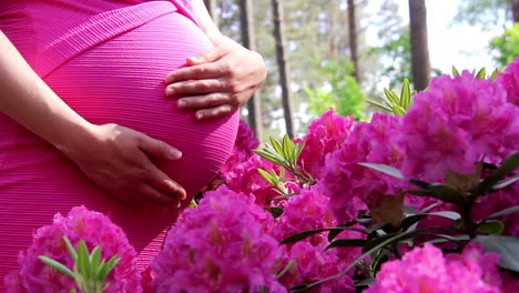 Pregnant-woman-in-dress-holds-hands-on-belly-on-natural-background-of-rhododendron-at-summer-day
