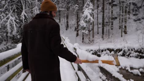 Young-East-european-men-with-brown-hat-and-black-jacket-drags-an-axe-over-a-snowy-railing-of-a-bridge
