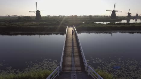 A-drone-shot-panning-up,-while-a-girl-walks-and-turns-around-on-a-bridge,-looking-at-Dutch-Windmills-in-the-Netherlands-during-sunrise