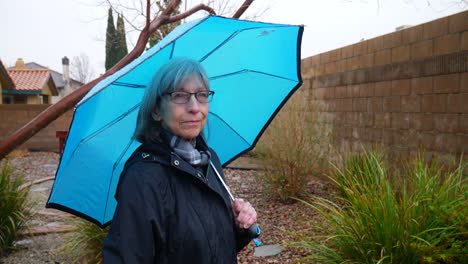 A-beautiful-old-woman-walking-and-enjoying-her-backyard-garden-with-a-blue-umbrella-in-the-rain-during-a-winter-storm