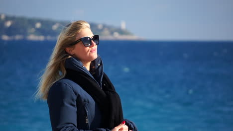 A-young-woman-waves-her-blond-hair-in-the-wind,-the-blue-sea-in-soft-focus-behind-her-on-a-sunny,-windy-day-SLOMO