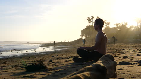 A-man-sits-in-a-meditating-pose-for-stress-relief-on-a-sunset-beach-with-palm-trees-and-people-in-silhouette-on-the-ocean-shore
