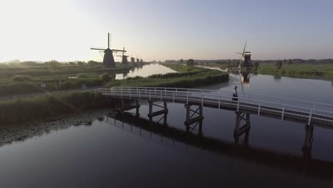 A-tripod-drone-shot,-filming-a-model-walking-over-a-bridge,-with-Dutch-Windmills-in-the-background-during-sunrise