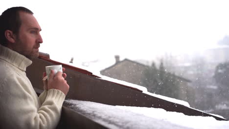 Mann-Trinkt-Tee-Und-Spielt-Mit-Schneeflocken-Auf-Einem-Balkon,-Während-Es-Schneit