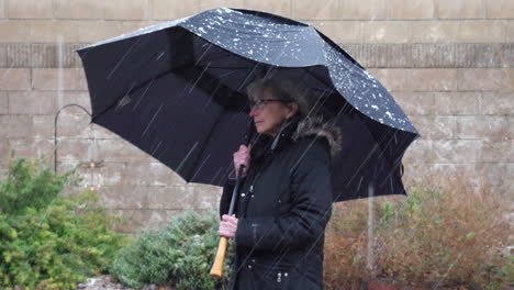 Eine-Alte-Frau-Mit-Schwarzem-Regenschirm-Und-Regenmantel-In-Einem-Eiskalten-Wintersturm-Bei-Schlechtem-Wetter,-Während-Schnee-Und-Hagel-Um-Ihre-Zeitlupe-Fallen