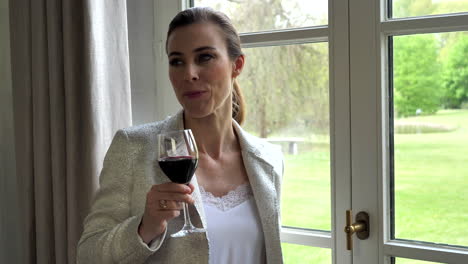 Classy-mature-adult-woman-sipping-red-wine-looking-through-window,-view-on-beautiful-garden,-slow-motion