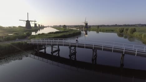 A-drone-shot-panning-left,-around-a-girl-walking-and-turning-around-on-a-bridge,-looking-at-Dutch-Windmills-in-the-Netherlands-during-sunrise