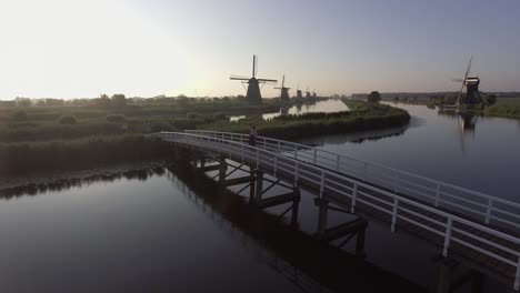 Eine-Drohne-Schwenkt-Nach-Links,-Um-Ein-Modell-Herum,-Das-Auf-Einer-Brücke-Steht-Und-Bei-Sonnenaufgang-Auf-Holländische-Windmühlen-In-Den-Niederlanden-Blickt