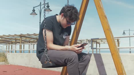 Young-man-sitting-on-swing-in-playground-on-phone