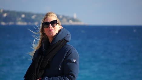 A-young-blonde-woman-laughs-to-the-camera-with-the-blue-sea-in-soft-focus-behind-her-on-a-windy,-sunny-day-SLOMO