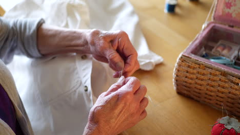 The-wrinkled-hands-of-an-aging-old-woman-untangling-a-knot-in-thread-while-repairing-a-button-on-a-white-dress-shirt-next-to-her-sewing-kit