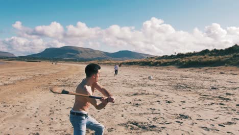 Young-Man-Tossing-and-Hitting-a-Hurley-Ball-with-a-Hurley-Stick-on-the-Beach-with-a-Beautiful-Mountain-Landscape-in-Background