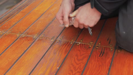 Young-man-uses-pocket-knife-to-clean-out-old-mastic-of-wooden-boat-roof-planking-close-up
