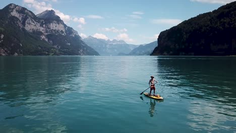 A-white-male-is-holding-up-his-paddle-on-a-stand-up-paddle,-sup,-the-drone-flies-up-to-show-him-in-the-middle-of-a-lake-in-Switzerland