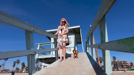 Beautiful-young-blonde-girl-and-cute-dog-walk-down-a-lifeguard-station-ramp-on-Venice-Beach-in-slow-motion