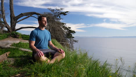 A-fit-young-caucasian-man-sitting-down-to-practice-meditation-in-and-mindfulness-in-nature-on-a-cliff-over-the-blue-ocean-and-beach-of-Santa-Barbara,-California