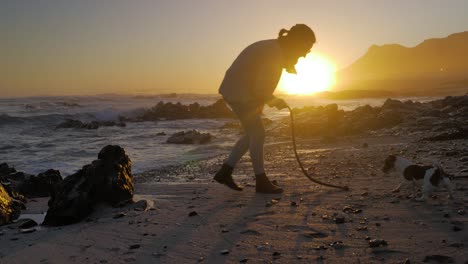 Young-woman-plays-with-Jack-Russell-dog-on-beach-during-gorgeous-sunset,-small-waves-crashing-onto-rocky-beach