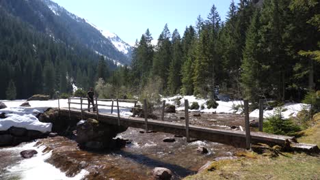 Still-shot-of-a-young-man-walking-over-a-wooden-bridge-on-a-hiking-route-in-the-Swiss-alps-over-a-calm-river