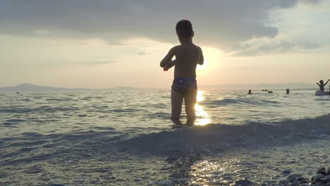 Kind-Spielt-An-Den-Wellen-Mit-Sonnenuntergang-Im-Hintergrund-Am-Strand-Von-Kalamata,-Griechenland