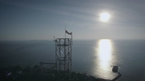 Aerial-shot-of-an-BTS-Base-Transceiver-Station-steel-tower-in-a-small-island,-backed-with-sunset,-surrounded-by-blue-ocean-in-the-afternoon
