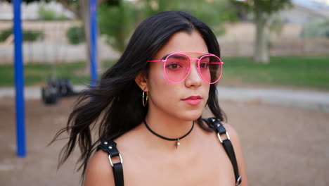 Eine-Junge-Hispanische-Hipster-Frau,-Die-Vintage-Modekleidung-Und-Retro-Pink-Fliegersonnenbrille-Auf-Einem-Parkspielplatz-Trägt