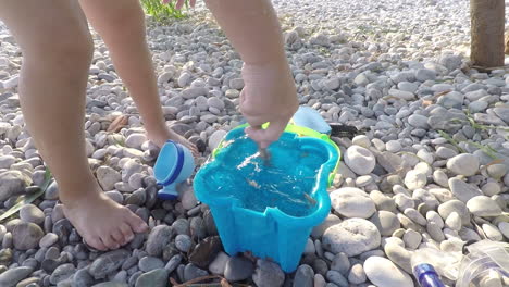 Toddler-stirs-sea-water-in-a-blue-toy-bucket-with-a-branch