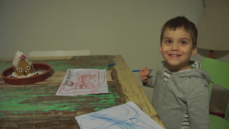 Tilt-shot-of-Caucasian-toddler-boy-drawing-with-colour-markers-on-paper,-and-smiling-at-the-camera
