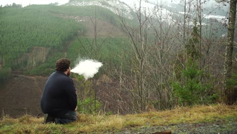 A-young-Millennial-man-kneeling-down-and-using-a-vape-machine-and-blowing-smoke-while-enjoying-being-outdoors-in-nature,-slow-motion