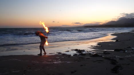 A-man-in-silhouette-spinning-a-fire-staff-on-the-beach-at-sunset-with-flames-and-ocean-waves-in-slow-motion