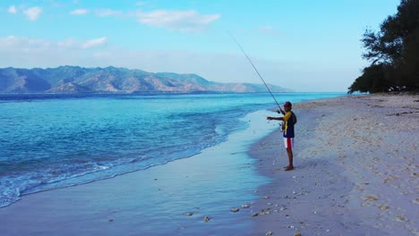 Junger-Mann-Fischt-Eine-Kleine-Fischrolle-Aus-Dem-Blauen-Meer-Am-Leeren-Strand-Bei-Sonnenuntergang-Bei-Schwachem-Licht,-Bali
