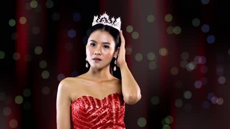 Schönheitskönigin-Mit-Asiatischen-Gesichtszügen-Und-Rotem-Kleid-Lächelt-Und-Posiert,-Während-Sie-Ihre-Krone-Trägt---Bühnenvorhanghintergrund-Mit-Bokeh