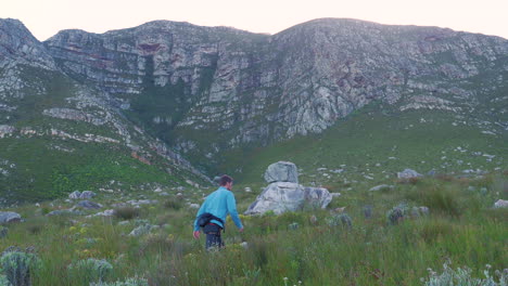 Man-hiking-up-trail-next-to-imposing-sandstone-mountains,-Hermanus,-South-Africa