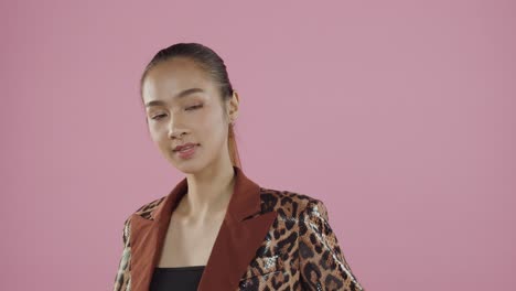 Attractive,-young-Asian-woman-in-fashionable-leopard-print-dress-poses-with-a-pretty-smile-and-hair-pulled-back-from-her-face-in-a-ponytail---professional-studio-with-pink-backdrop