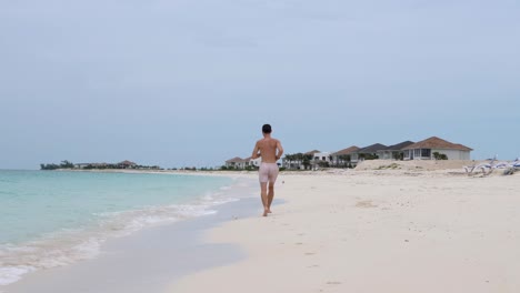 Young-topless-white-male-runs-on-the-shore-of-the-beach-in-Bimini,-Bahamas-along-some-new-villas