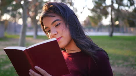Close-up-on-a-young-hispanic-woman-college-student-reading-the-pages-of-a-red-story-book-or-novel-outdoors-at-sunset-SLOW-MOTION