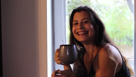Happy-beautiful-woman-with-a-cup-of-coffee-sitting-on-window-sill