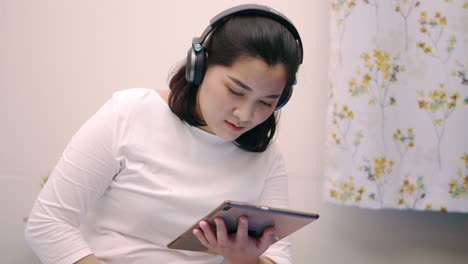 Cheerful-young-beautiful-Asian-woman-relaxing-and-listening-to-music-by-tablet-with-headphones-on-the-bed-at-home-lifestyle-and-pleasure-concept