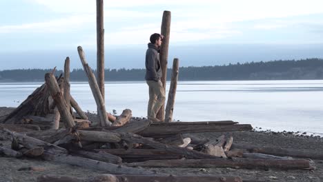 Man-admiring-the-stunning-landscape-view,-stood-on-drift-wood-on-the-coast-of-Vancouver-Island,-Canada