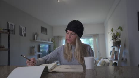 A-beautiful-blonde-woman-writing-on-a-notebook-and-drinking-a-hot-beverage-at-home