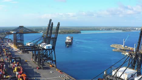 Scenic-flight-above-cargo-shipping-container-sea-port,-cranes-towards-cargo-ship-pulled-by-tugboat,-Punta-Caucedo,-Dominican-Republic,-overhead-aerial-approach
