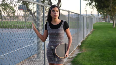 A-pretty-young-hispanic-woman-walking-through-an-urban-city-park-with-her-skateboard-along-a-chainlink-fence-SLOW-MOTION