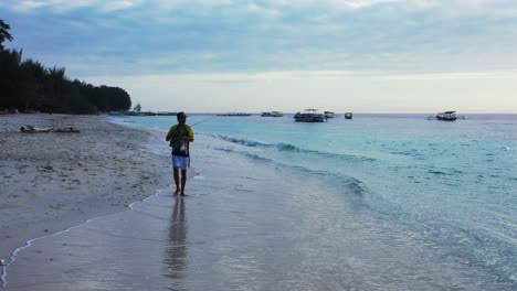 Pescador-Caminando-Por-La-Playa-De-Arena,-Sosteniendo-Una-Caña-De-Pescar-Lista-Para-Lanzar-La-Línea-Al-Mar,-Observando-Cuidadosamente-En-La-Laguna-Señales-De-Peces-En-El-Mar-Indonesio
