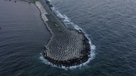 Scenic-top-view-of-long-stone-rocky-jetty-in-blue-ocean-sea-water-and-waves-crashing-onshore,-above-circle-aerial