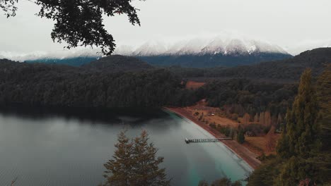 View-from-a-viewpoint-to-the-shore-of-a-lake-with-a-pier,-forest-and-snowy-mountains-in-the-background