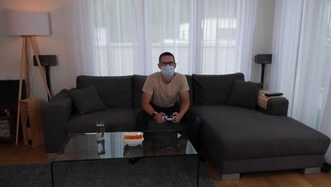Young-man-with-face-mask-sitting-on-sofa-playing-video-games