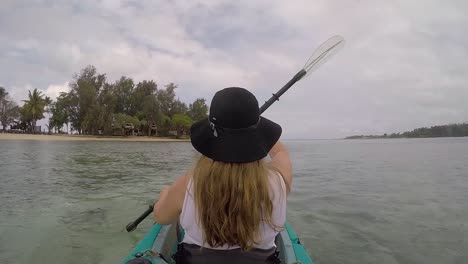 Girl-with-long-blonde-hair-wearing-a-black-hat-paddling-a-kayak-near-a-tropical-island