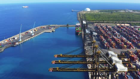 Colorful-cargo-containers-stacked-at-shipyard-terminal-by-tall-cranes-with-view-of-vast-blue-ocean-sea-water,-Punta-Caucedo,-Dominican-Republic,-aerial
