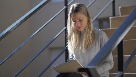 A-joyful-blonde-woman-sitting-on-the-stairs-and-writing-on-a-notebook